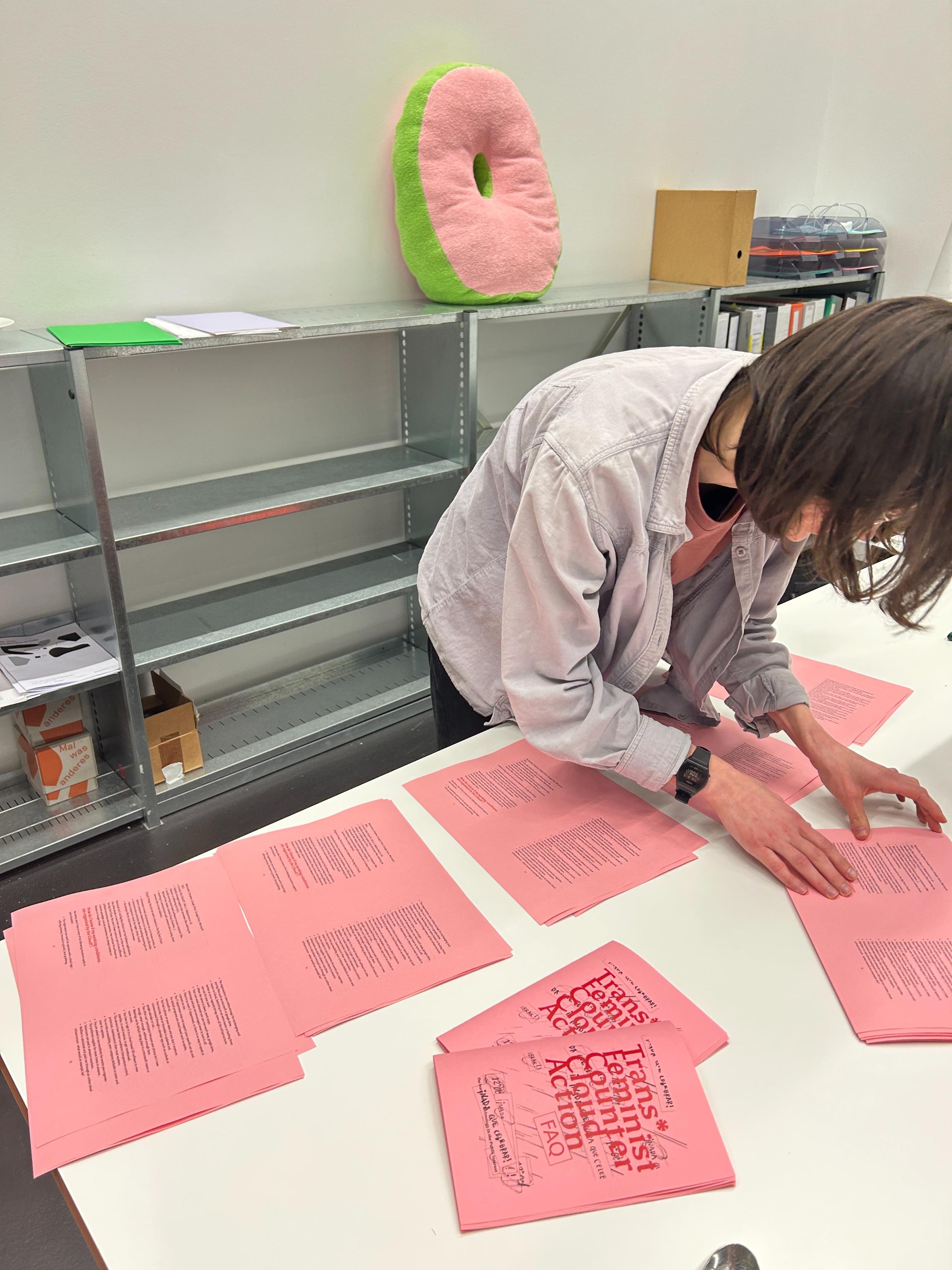 Cristina is bending over a table to organize freshly printed bright pink pages of the "Trans* Feminist Counter Cloud Action FAQ” zine. The cover text of the zine is in red ink, and visible from the two copies that are folded on the table. A flamboyant love letter to trans*feminist life and flourishing beyond the cloud, it draws you in and makes no apologies. Cassandra is taking the picture at Critical Media Lab in Basel, and helping to fold copies with the spoon that is just barely visible in the image at the bottom lefthand corner, to bring to the gathering in Grenoble, France hosted by STopMicro Collective. It’s late in the evening, and Cristina is preparing to take the night train back to Rotterdam and bring copies with her, there’s an assortment of snacks and drinks outside the shot. Lit with strong fluorescent lighting, behind Cristina is a large O-shaped pillow (made by MA Transversal Design graduate Nani) on a steel utilitarian bookshelf, one letter that helps to spell out "ABOLITION" when they all come together. The pink face of the pillow matches the pink of the page, as they reverberate in conversation. Pleasurable in pink, terrycloth plush of the stuffed letter absorbing light and color that bounces off the clean white wall into the neon green fabric that holds the two sides of the O together. All the letters are meant to be used, but they’re delicate as well, sewed by hand. They bring us back to a different form of clouding, of being grounded in careful attention to the daily work of abolition. Your head can rest on a cloud beyond the cloud, while your feet remain firmly on the ground. (Insert or link to image of sticker made by Femke). The other letters are mostly gathered in the front of the office in a space of couches and chairs meant for conversation, reading or lounging. A playful, rejuvenating reminder of the everyday practices we embody, what bolsters or support us, and the places and ways we make collective life outside of the private home. The spaces we make for life and comfort in sites where we’re told we should not. We insist upon it regardless.