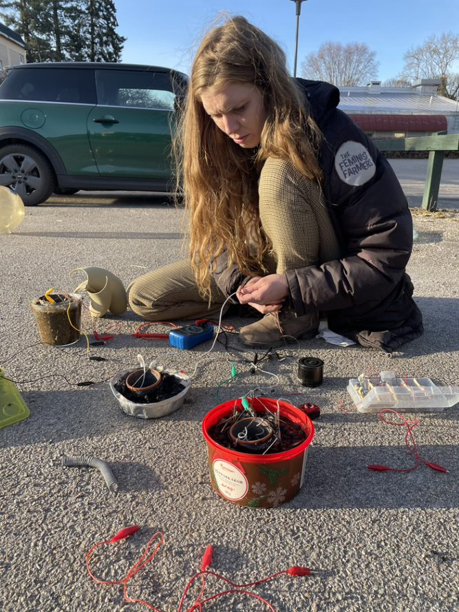 A person with long hair, wearing a jacket with a Feminist Farmers badge sits on the ground in a car park connecting fuel cells, which are housed in random containers