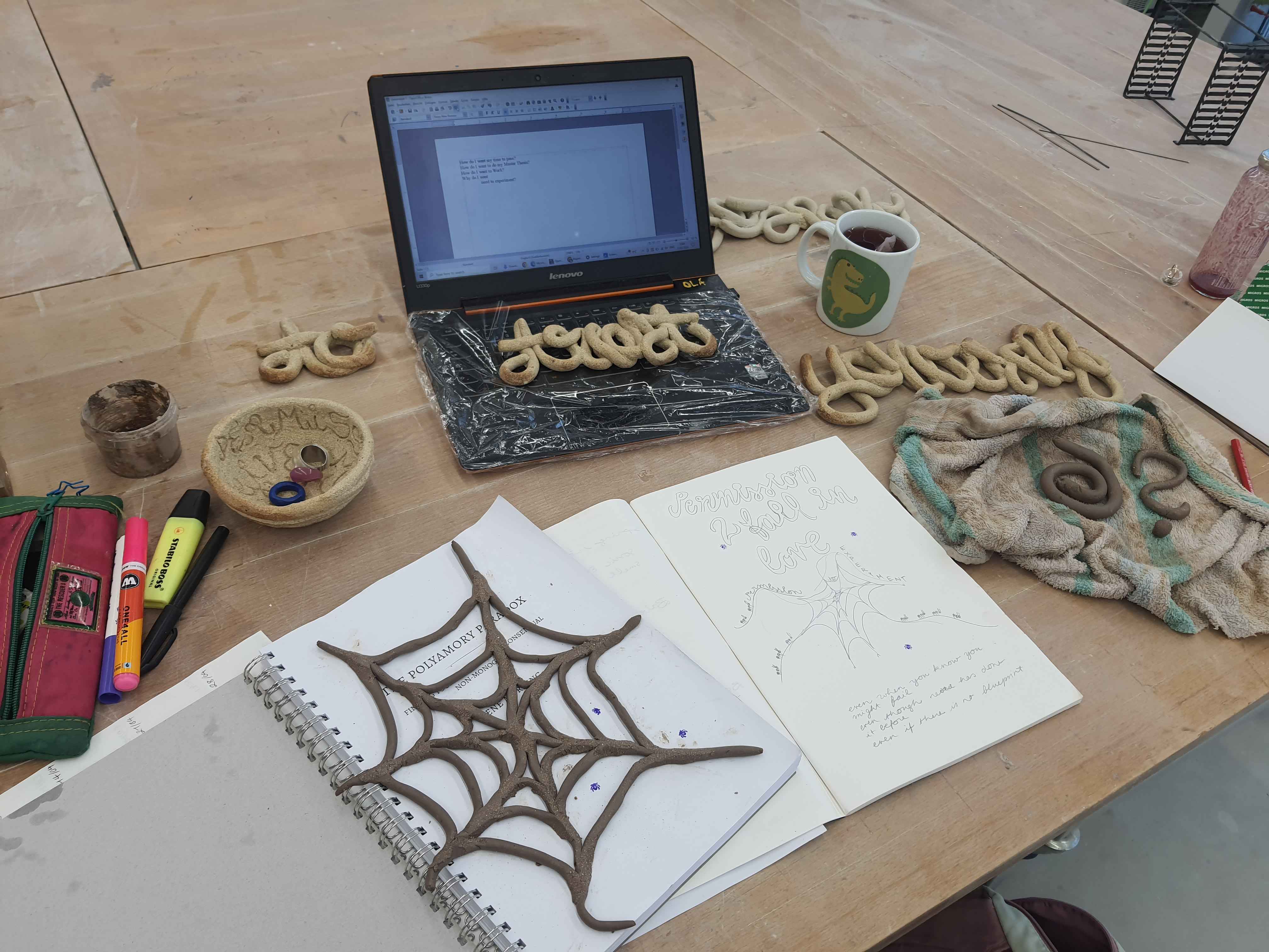 A picture of the table I was working on in the schools ceramic studio. There is the copy of polyamory paradox with a freshly coiled web on it. There is my computer where im writing passages from the book into, with the burnt coiled encrypted "trust"on it, the "yourself" is laying next to it. There is a frshly brewed cup of tea. There is a freshly coiled spiral and questionmark laying on a damp towel. There is my notebook where i wrote myself a "permission to fall in love" but turned into a "PErmission to fail in love". underneath is is a drawing of a spidernet, with little ants walking around it.