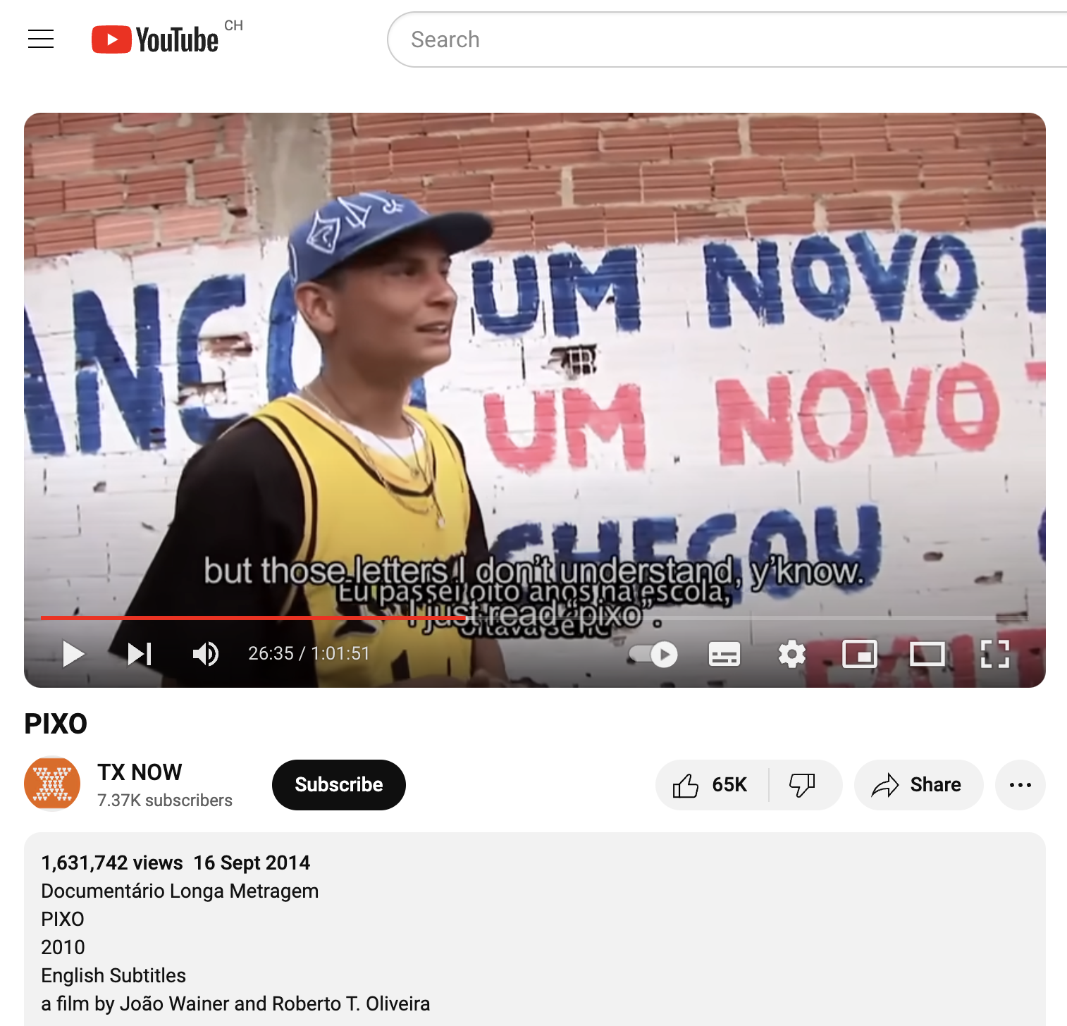 Its a screenshot from the Youtube page of the documentary. There Is a young cic-man, wearing a blue cap with white Pixo letters on it. He is wearing a black shirt and a yellow Top on top. He stand in front of a painted brick wall. At the top you can see the bare, orange bricks and underneath you can read parts of a text in (what he calls) "straight letters" (not pico) "...um novo, um novo chegou" . There is an over lap of subtitles. in portuguese it says: "Eu passei oiro anos na escola, oitava serie" over it in english it says: "but those letters I dont understand, y'know. I just reas "pixo".