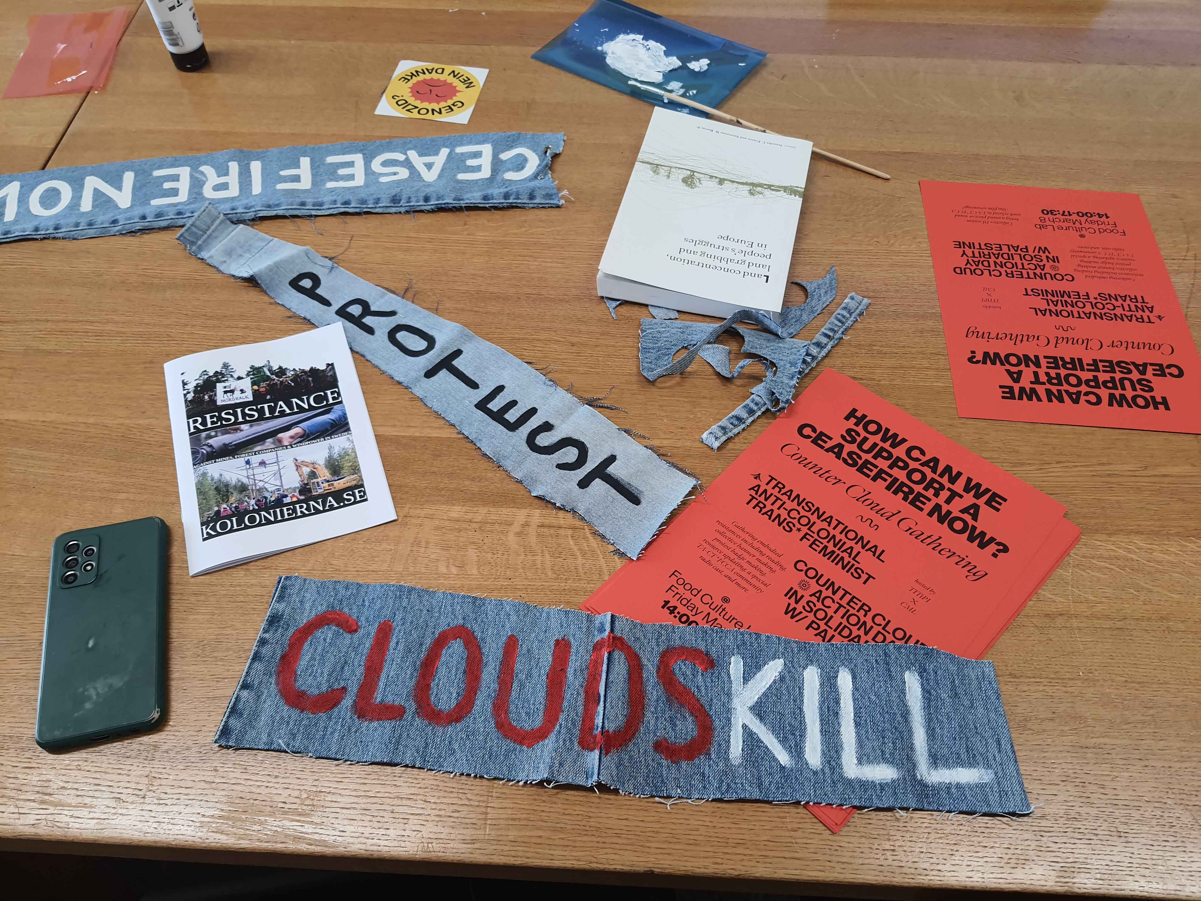 Picture of a Table,There are the Flyers for the Counter Cloud Gathering Event on the 8M where the workshop was held together with a Radio Show, at the Food Culture(s) Lab in BAsel, With Titipi. there are Various Jeans Scraps with Writing on it, the say "Ceasfire now!", "Protest" and "Clouds Kill", there is a zine laying It says "Resistance Kolonierna.SE", A Sticker that says "Genozid? Nein Danke" with an unhappy sun, and a book titeles "Land concentration, land grabbing and peoples struggles in Europe"