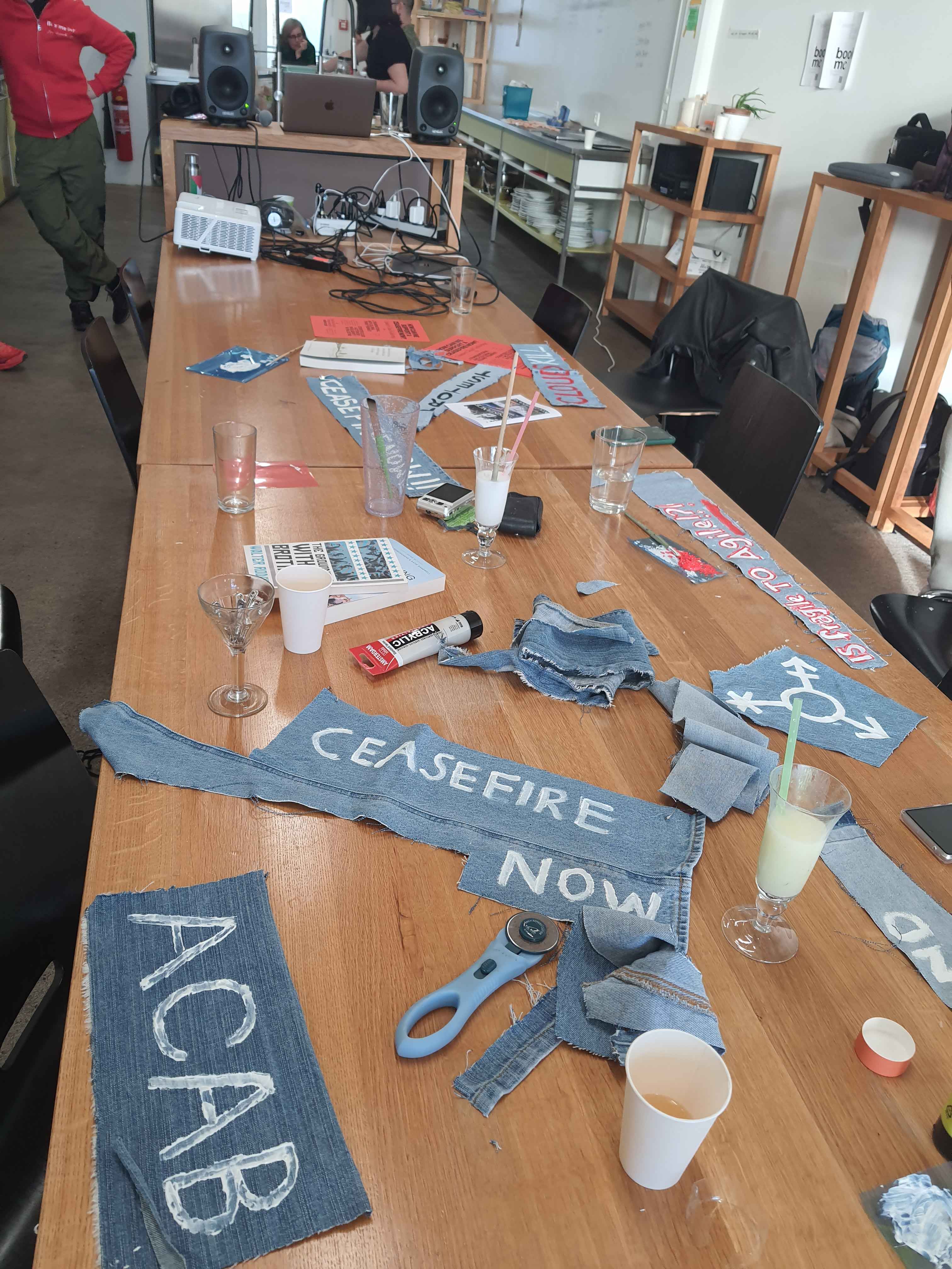 Picture of a Table, for the Counter Cloud Gathering Event on the 8M where the workshop was held together with a Radio Show, at the Food Culture(s) Lab in Basel, With Titipi. There are Various Jeans Scraps with Protest Text on it,there is paints, brushes,zines and Books laying around. At the end of the Table there is the Radio set up.