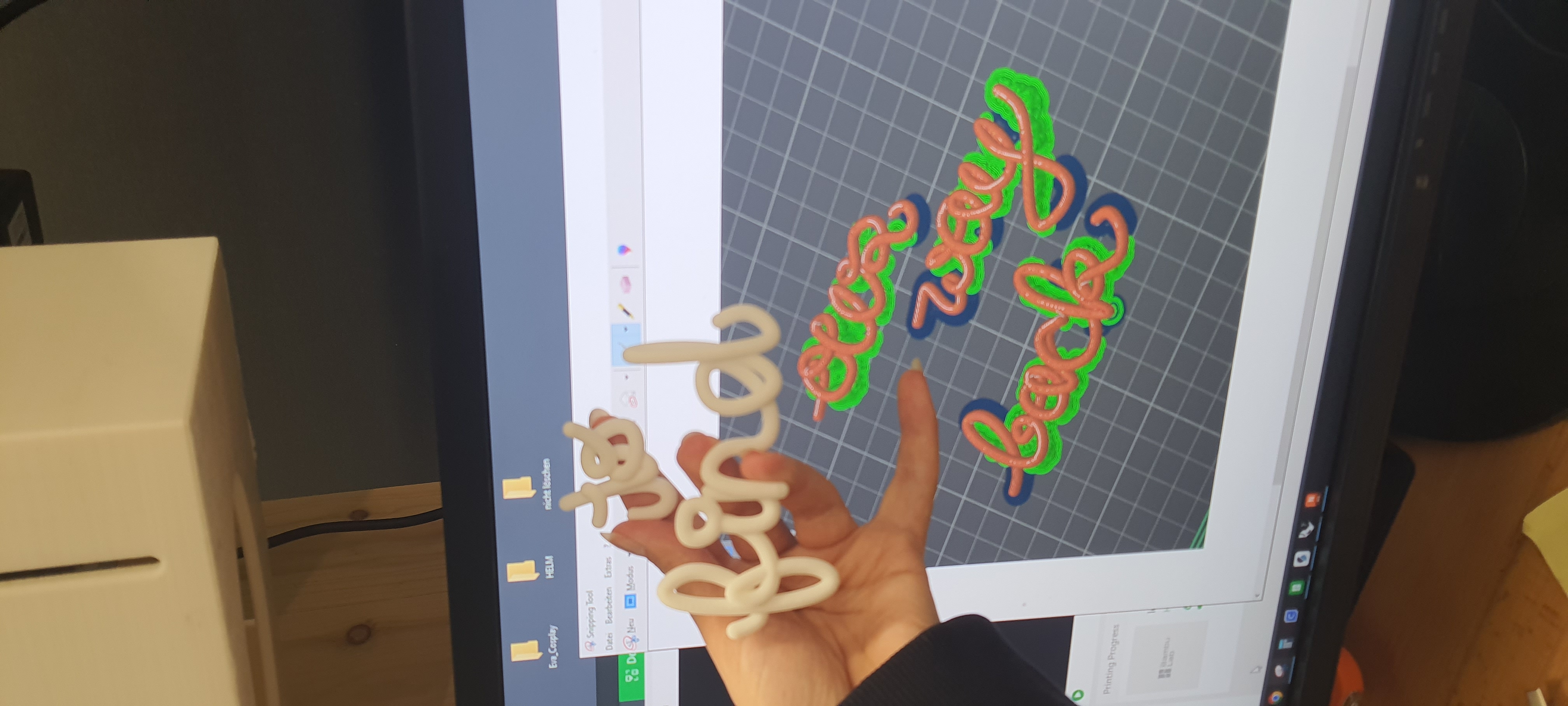 A picture of a hand holding two 3d printed words in front of a screen of two new words being prepared to print.