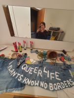 A mirror selfie, I'm feeling happy, a chaotic working table, a toolbelt in the making, Pieces of cut Jeans, Acrylic paint tubes, brushes, tools (mini basket, rubber fish, rope, cherries, chrystals) laying around, white acrylic on Denim, saying in big QUEER LIFE, underneath in smaller: PLANTS KNOW NO BORDERS, you could also read QUEER LIFE KNOWS NO BORDERS.