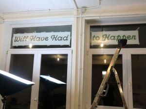 glass door with the lettering "Will Have Had to Happen", shot taken from a white-walled indoors towards a dark outdoors